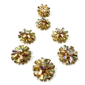 Può includere: Una collezione di gioielli vintage dorati a forma di stella, con accenti di cristalli iridescenti e sfaccettati. La composizione include diversi orecchini e un bottone, tutti con un aspetto radioso e scintillante.