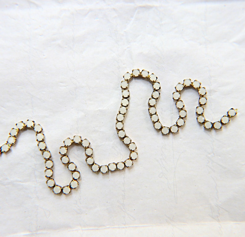 Puede incluir: Un delicado collar de cadena dorada con una serie de peque&ntilde;as piedras preciosas blancas y redondas. El collar est&aacute; dispuesto en un patr&oacute;n ondulado sobre un fondo blanco texturizado, creando un dise&ntilde;o simple pero elegante.