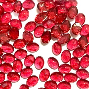 May include: A collection of numerous small, oval-shaped red glass cabochons scattered across a white surface. Each piece features a smooth, rounded top with a translucent, deep red colour and subtle internal swirling patterns throughout the glass.