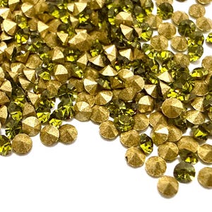 May include: A collection of small, round, faceted embellishments in olive green and metallic gold. Each piece features a pointed, conical back and a flat, multi-faceted top surface that reflects light, creating a textured, scattered arrangement.