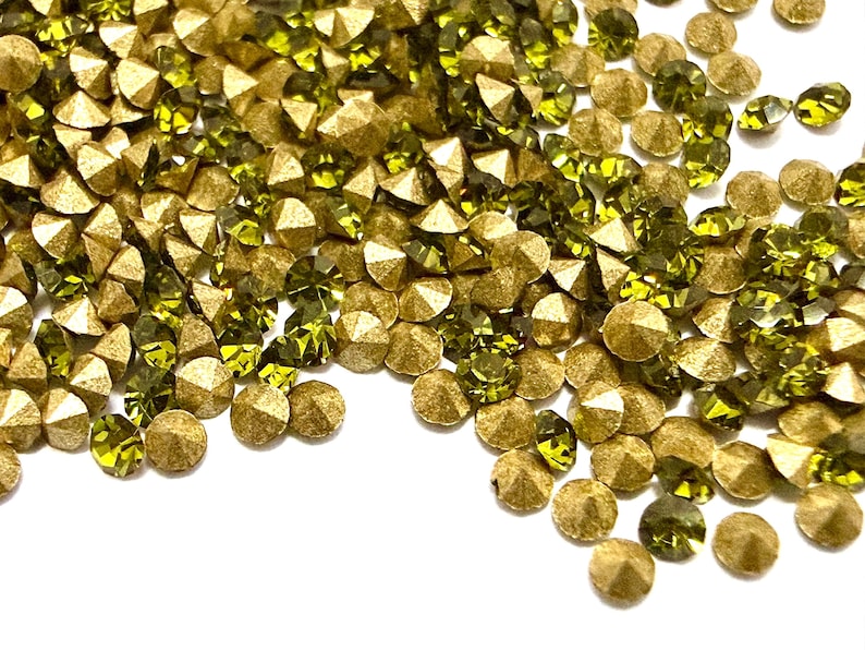 May include: A collection of small, round, faceted embellishments in olive green and metallic gold. Each piece features a pointed, conical back and a flat, multi-faceted top surface that reflects light, creating a textured, scattered arrangement.