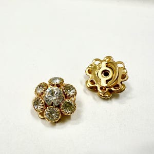 Può includere: Due orecchini dorati a forma di fiore con strass chiari e scintillanti. Ogni orecchino presenta uno strass centrale più grande circondato da pietre più piccole. Il retro di un orecchino è visibile, mostrando il perno e il meccanismo di chiusura.