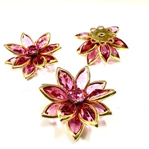 May include: Three flower-shaped brooches with pink crystal petals and a gold-colored metal frame. Each brooch has a central pink crystal. The brooches are arranged on a white background.