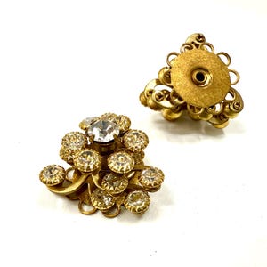Può includere: Due orecchini a clip vintage in metallo dorato. Un orecchino presenta un gruppo di strass rotondi e trasparenti e strass più piccoli color ambra. L'altro orecchino ha un design simile, con un retro rotondo e filigrana dorata decorativa.