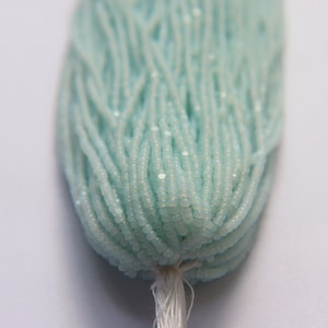May include: A strand of light blue seed beads, also known as rocailles, with a white iridescent finish. The beads are strung together and tied off with a white thread.