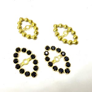 Può includere: Quattro elementi decorativi per gioielli. Due sono dorati con una serie di piccole protuberanze rotonde sul bordo. Gli altri due sono dorati con pietre preziose nere e rotonde sul bordo. Tutti hanno un piccolo anello per il fissaggio.