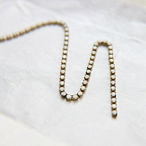 Puede incluir: Un delicado collar con peque&ntilde;as piedras preciosas redondas y transparentes engastadas en un metal dorado. El collar est&aacute; dispuesto en un patr&oacute;n ondulado sobre un fondo blanco texturizado. Las piedras preciosas capturan la luz, creando un brillo sutil.