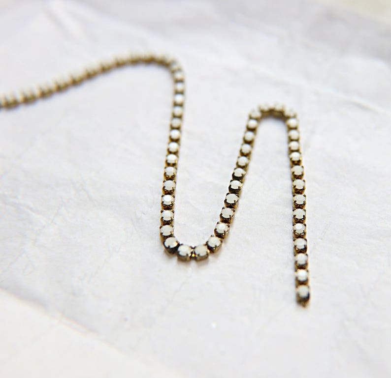 Puede incluir: Un delicado collar con peque&ntilde;as piedras preciosas redondas y transparentes engastadas en un metal dorado. El collar est&aacute; dispuesto en un patr&oacute;n ondulado sobre un fondo blanco texturizado. Las piedras preciosas capturan la luz, creando un brillo sutil.