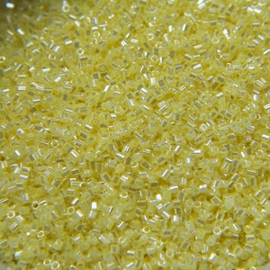 May include: A close-up image of a large quantity of small, yellow, glass seed beads. The beads are shiny and have a slight iridescent quality.