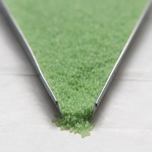 May include: A metal funnel filled with small, light green glass beads. The beads are spilling out of the funnel onto a white surface.