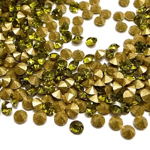 May include: A collection of small, round, faceted rhinestones in olive green and metallic gold. Each piece features a pointed, conical back and a flat, multi-faceted top surface that reflects light, creating a scattered, textured arrangement.