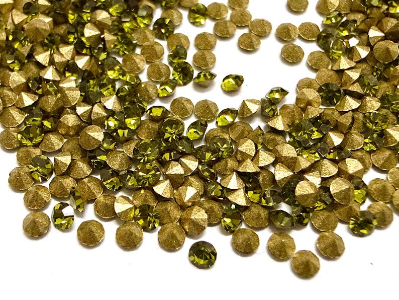 May include: A collection of small, round, faceted rhinestones in olive green and metallic gold. Each piece features a pointed, conical back and a flat, multi-faceted top surface that reflects light, creating a scattered, textured arrangement.