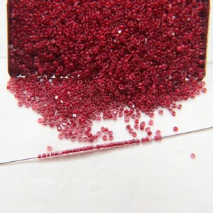 Puede incluir: Primer plano de un montón de pequeñas cuentas de vidrio rojas. Las cuentas están esparcidas sobre una superficie blanca, con algunas enhebradas en un alambre delgado.