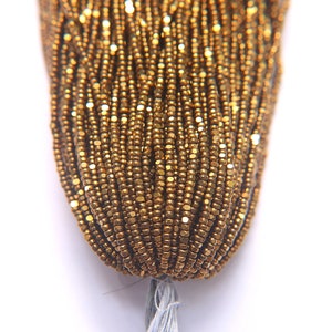 Puede incluir: Primer plano de una hebra de cuentas de semillas doradas. Las cuentas son peque&ntilde;as y tienen un brillo met&aacute;lico.