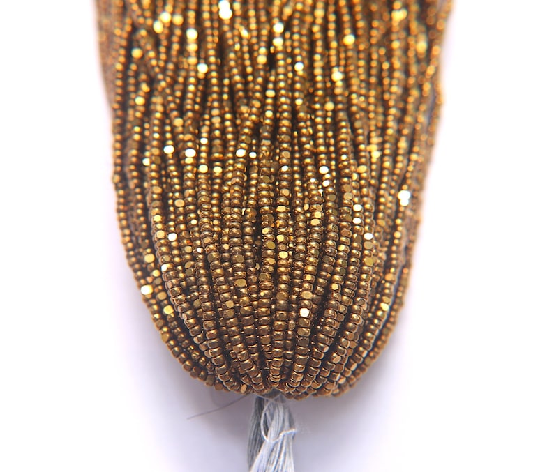 Puede incluir: Primer plano de una hebra de cuentas de semillas doradas. Las cuentas son peque&ntilde;as y tienen un brillo met&aacute;lico.
