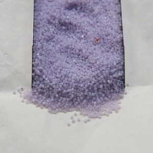 May include: A close-up of a metal scoop filled with small, light purple beads. The beads are spilling out of the scoop onto a white surface.
