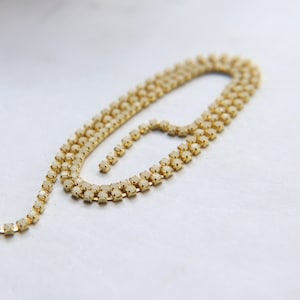 May include: A delicate gold-toned necklace with multiple rows of small, sparkling crystals. The necklace is arranged in a flowing, curved shape, showcasing the intricate design and the light-catching qualities of the crystals. The piece is set against a soft, neutral background.