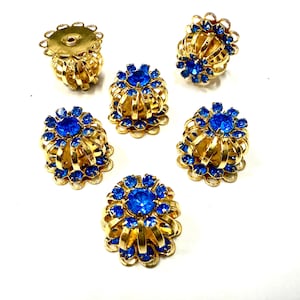 May include: Six vintage gold-tone clip-on earrings with vibrant blue rhinestones. Each earring features a central blue stone surrounded by smaller blue stones, set in a decorative gold filigree design. The earrings have a classic, elegant style.