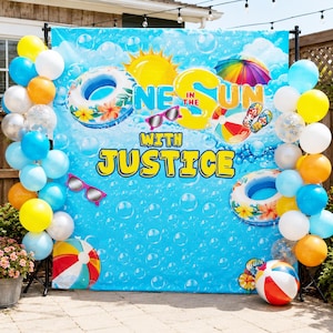 May include: A blue backdrop featuring a bubble pattern, a yellow sun, a beach umbrella, sunglasses, and two floral swim rings. The text reads ONE IN THE SUN WITH JUSTICE. The display is framed by a balloon arch in shades of blue, yellow, and white.