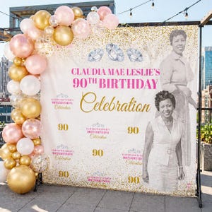 May include: A backdrop for a 90th birthday celebration, featuring a photo of a woman, the text "Claudia Mae Leslie's 90th Birthday Celebration," and diamond illustrations. A balloon arch in gold, pink, and white decorates the left side.