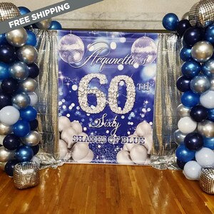 May include: A celebratory backdrop with a blue theme, featuring a banner that reads "Acqunetta's 60th Sixty Shades of Blue." The banner is flanked by columns of blue, silver, and white balloons. Disco balls and silver curtains enhance the festive scene.