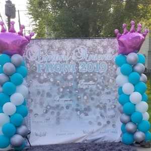 May include: A white and silver backdrop with the text "Amoni Amene's PROM 2019" surrounded by silver and blue balloons with a purple crown balloon on each side.
