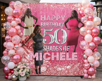 Op de afbeelding: Een roze achtergrond met de tekst "HAPPY BIRTHDAY 50 SHADES OF MICHELE" is omgeven door roze en witte ballonnen. Drie vrouwen poseren voor de achtergrond. Een bloemstuk en zilveren bollen staan op de voorgrond.