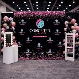 May include: A trade show booth for Conceited Cosmetics features a black backdrop with pink glitter and the brand's logo. Displayed are makeup products, a lighted mirror, and balloons in pink, black, and silver. A white shelving unit holds more products.