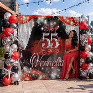 May include: A red and black birthday backdrop with the text "Happy 55th Birthday Vernetta" is surrounded by red, silver, and black balloons. A woman in a red dress sits in front of the backdrop.