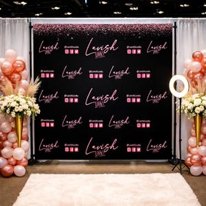 May include: A black backdrop featuring the word "Lavish" in pink script, repeated with social media icons. The backdrop is decorated with pink glitter. Balloon arrangements in pink and white, and floral arrangements flank the backdrop.
