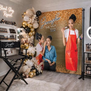 May include: A makeup studio scene featuring a backdrop with the text "Juresha The MUA." A makeup artist in a red apron holds a makeup brush and palette. A director's chair with the same logo is in the foreground, with shelves of makeup products.