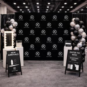Könnte beinhalten: Ein Messestand mit schwarzem Hintergrund und sich wiederholendem Logo. Zwei Werbetafeln zeigen "Neuheiten" und "Gewinnen Sie! Shopping Spree." Schwarze und silberne Ballons umrahmen die Ausstellung, mit einem beleuchteten Spiegel.