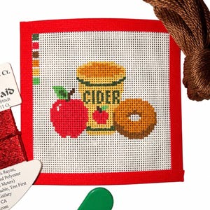 May include: A square needlepoint canvas with a red border featuring a cross-stitch design of a red apple, a yellow mug labeled CIDER, and a brown donut. Included are a skein of brown embroidery floss, a red thread card, and a green needle threader.