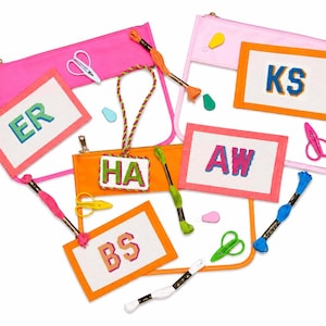 May include: A collection of clear vinyl pouches with neon pink and orange trim, featuring rectangular needlepoint canvases with embroidered letters ER, KS, AW, HA, and BS. Includes colorful embroidery floss skeins and small metal scissors.