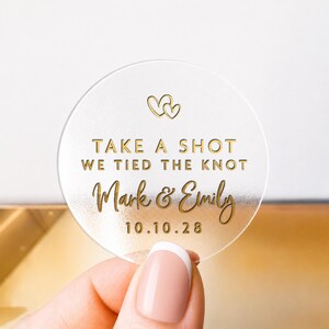 Pode incluir: Um autocolante redondo transparente com letras douradas e dois corações entrelaçados. O texto diz "TAKE A SHOT WE TIED THE KNOT" com os nomes "Mark & Emily" e a data "10.10.28".