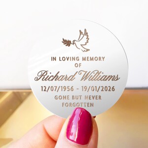 May include: A round, white memorial plaque with gold text and a dove illustration. The text reads "IN LOVING MEMORY OF Richard Williams 12/07/1956 - 19/01/2026 GONE BUT NEVER FORGOTTEN."