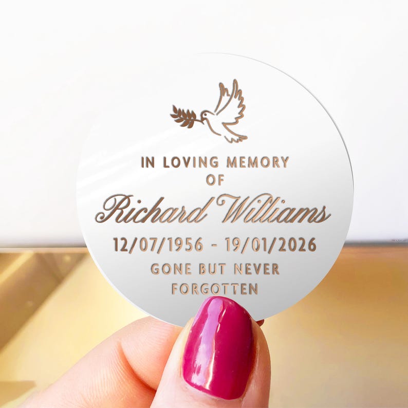 May include: A round, white memorial plaque with gold text and a dove illustration. The text reads "IN LOVING MEMORY OF Richard Williams 12/07/1956 - 19/01/2026 GONE BUT NEVER FORGOTTEN."