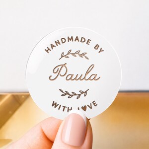 May include: A circular white sticker featuring copper-colored text and graphics. The text reads HANDMADE BY Paula WITH LOVE, with a small heart replacing the O in LOVE. Two stylized leafy branch illustrations are positioned above and below the name.