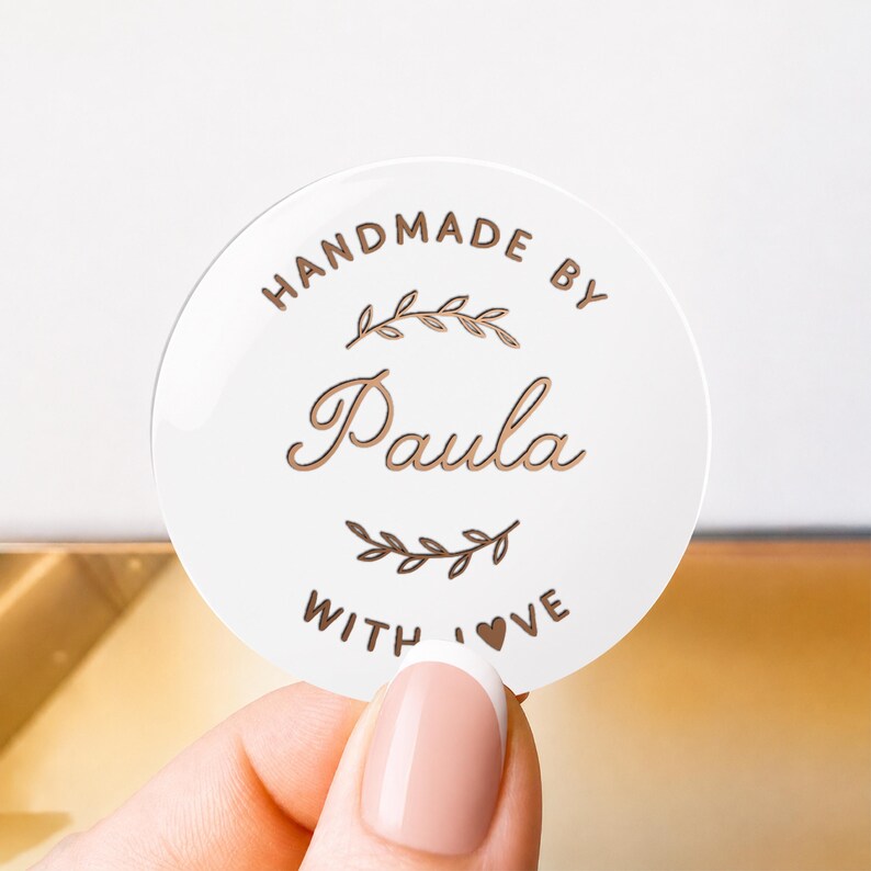 May include: A circular white sticker featuring copper-colored text and graphics. The text reads HANDMADE BY Paula WITH LOVE, with a small heart replacing the O in LOVE. Two stylized leafy branch illustrations are positioned above and below the name.