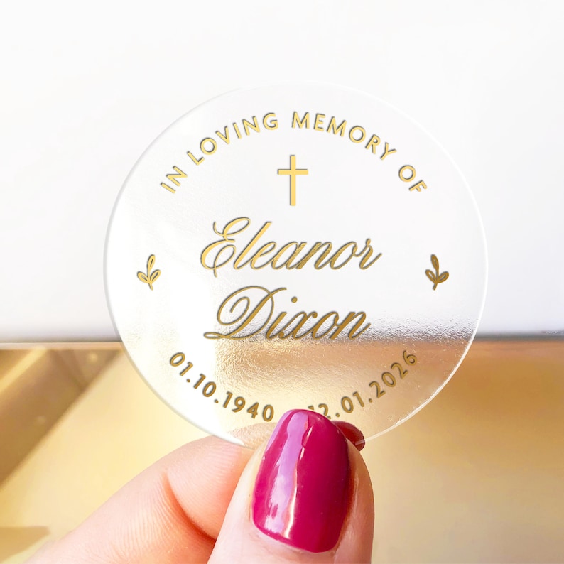 May include: A clear, round memorial sticker with gold lettering. The text reads "IN LOVING MEMORY OF Eleanor Dixon" with a cross and dates 01.10.1940 - 12.01.2026. Two small leaf designs flank the text.