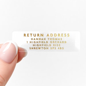 May include: A clear, rectangular return address label with gold lettering. The text reads "RETURN ADDRESS" above the name "HANNAH THOMAS" and an address. The label is held by a person's hand.