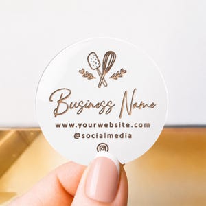 Pu&ograve; includere: Un'etichetta circolare in acrilico bianco con dettagli metallici color oro rosa. Il design include un'icona di una spatola e una frusta, il testo Business Name, un URL di sito web, un handle di social media e un'icona di Instagram.