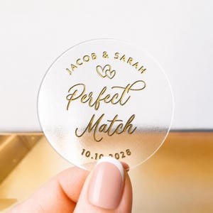 May include: A transparent, circular sticker with gold text. It features the names "Jacob & Sarah", two hearts, and the words "Perfect Match". The date "10.10.2028" is also present. The sticker is held by a hand with a French manicure.