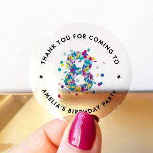May include: A clear, circular sticker with a gold foil backing. The sticker has the text "THANK YOU FOR COMING TO AMELIA'S BIRTHDAY PARTY" in black, surrounding a colourful number 8 made of confetti. Two small black stars are also present.