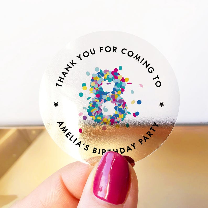 May include: A clear, circular sticker with a gold foil backing. The sticker has the text "THANK YOU FOR COMING TO AMELIA'S BIRTHDAY PARTY" in black, surrounding a colourful number 8 made of confetti. Two small black stars are also present.