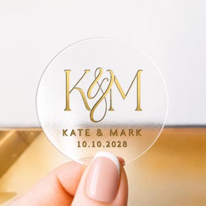 Könnte beinhalten: Ein klarer, runder Aufkleber mit goldfarbenem Schriftzug. Der Aufkleber zeigt die Initialen "K&M" in einer stilisierten Schriftart, mit den Namen "KATE & MARK" und dem Datum "10.10.2028" darunter. Der Aufkleber wird von einer Person gehalten.