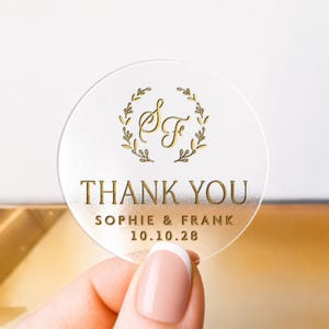 Pode incluir: Um autocolante redondo transparente com letras e design dourados. O texto "THANK YOU" é exibido em destaque, juntamente com os nomes "SOPHIE & FRANK" e a data "10.10.28". Uma grinalda floral dourada envolve as iniciais "S & F".