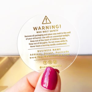 Op de afbeelding: Een ronde, transparante sticker met gouden tekst die "WAARSCHUWING! WAX MELT SAFETY" zegt en veiligheidsinstructies bevat voor het gebruik van wax melts. De sticker bevat ook de bedrijfsnaam, website en social media-handvatten.