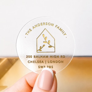 May include: A circular, frosted acrylic sticker featuring gold foil text and a house-shaped icon with a branch design. The text reads THE ANDERSON FAMILY, 200 BALHAM HIGH RD, CHELSEA | LONDON, and SW3 2BS, printed in a clean, sans-serif font.