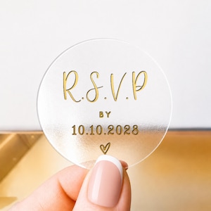 May include: A clear, circular sticker with gold text reading "R.S.V.P. BY 10.10.2028" and a heart symbol. The sticker is held by a person with a French manicure.